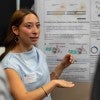 A young woman gestures at a poster while talking to an audience. 