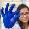 A person in blue gloves holds a tiny, patterned object, with a blurred background showing another individual in a lab setting.