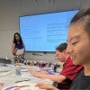 A smiling asian woman sits at a table; a woman behind her reads notes in front of a presentation 