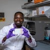 A black man in a lab coat and gloves holds up a 96 well plate while smiling 