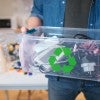 A white person holds a plastic bin full of electronic waste. 