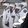 Four astronauts wearing white and black SpaceX spacesuits and white helmets walk together outdoors near a vehicle, with three waving toward the side.