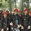 A diverse group of eight young people wearing safety helmets, harnesses, and protective gloves pose together outdoors on a platform surrounded by a lush forest. 